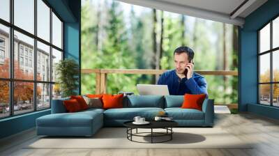 Serious middle aged man talking on cell phone and using laptop computer sitting at wooden table on country house balcony. Man working remotely online Wall mural