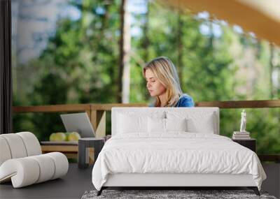 Side view of young woman working on laptop computer sitting at wooden table on balcony of countryside house Wall mural