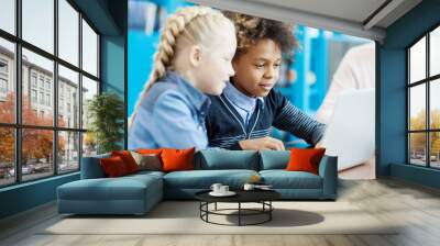 Two elementary school kids, mixed race boy and Caucasian girl, studying on laptop computer sitting at desk in classroom Wall mural