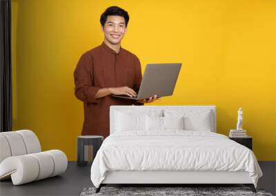 Asian business man holding a laptop computer and standing isolated on yellow background Wall mural