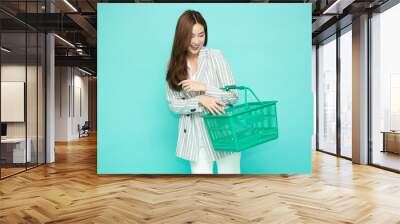 Beautiful Asian woman smiling and holding shopping basket isolated on light green background Wall mural