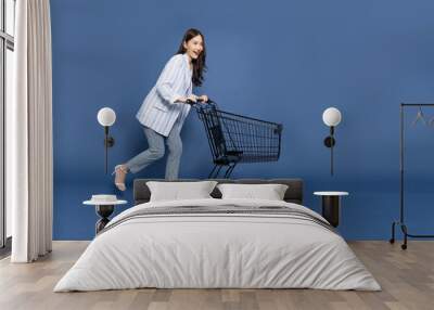 Full length portrait of young Asian woman pushing an empty shopping cart or shopping trolley isolated on deep blue background Wall mural