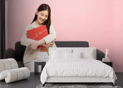 Happy beautiful Asian woman smile hugging red gift box and sitting on sofa isolated on pink background. In love, Receiving gifts from lovers. New Year or Christmas concept Wall mural