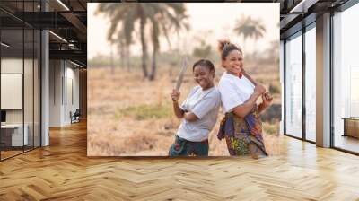 portrait of two happy female african farmers standing back to back, holding farm tools Wall mural