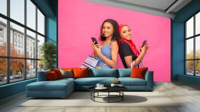 two gorgeous young african ladies holding shopping bags standing back to back, holding their phones, smiling Wall mural