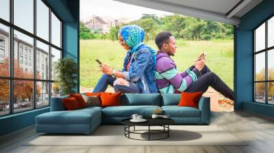 two young black people sitting back to back outside in a park, using their phones Wall mural