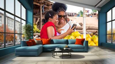 two young women in a local market, a trader and her customer, viewing content on a phone together Wall mural