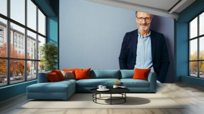 Confident middle-aged man in blue blazer and blue shirt, wearing glasses, standing against plain blue background with copy space and looking at camera Wall mural