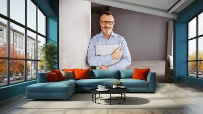 Older Businessman in Blue Shirt, Relaxed Leaning against a Column in the Office, Holding a Folder Wall mural