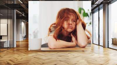 Red-haired girl sitting at table with boring face Wall mural