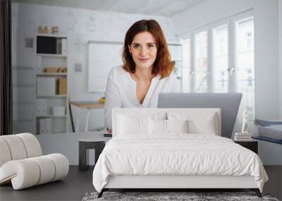 young woman sits in the office in front of her laptop and looks into the camera Wall mural