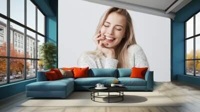 Beautiful female closing eyes because of joy, with long blonde hair and gentle smile. Cheerful woman holds hand under chin, has pleased expression of face. Face expression and positive emotions Wall mural