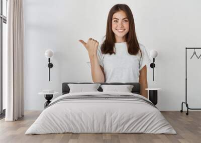 Beautiful female student making her choice and pointing thumb left, showing company logo or banner, standing over white background Wall mural