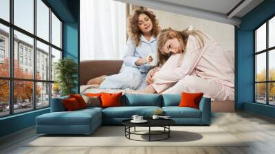 Best friend cheer up heartbroken blonde. Portrait of two attractive women sitting on sofa in nightwear. Curly-haired girl drinking coffee, patting sister on head while she is upset or feeling cramps Wall mural