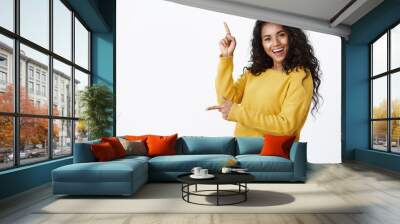 Cheerful carefree african-american woman in yellow sweater with curly haircut, smiling and laughing joyfully dancing as pointing sideways, showing left and up copy space, white background Wall mural