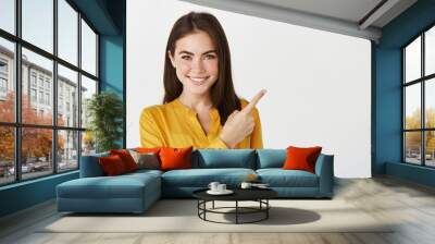 Close-up of brunette female model showing promo offer, pointing finger right and smiling at camera, white background Wall mural