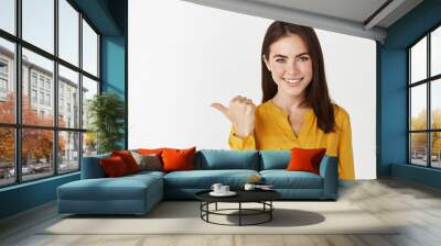 Close-up of confident and happy brunette girl pointing thumb left, checking out promo offer on white copy space Wall mural