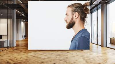 Close up of good-looking bearded hipster guy with hair in bun, in blue t-shirt standing in profile, looking aside, posing for photo. Wall mural