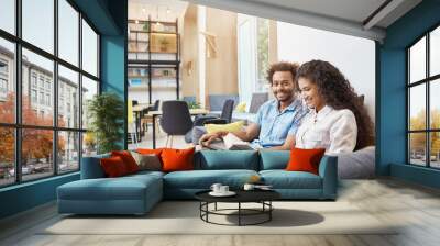 Close up of two young people in casual clothes sitting on sofa in modern library. Girl reading to book, getting ready for universit lecture. Guy looking in camera with happy face expression, holding Wall mural