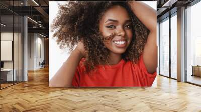 Close-up shot of carefree relaxed and tender african-american female model in red t-shirt playing with curly hair holding hands on head tilting and smiling caring with sensual gaze over white wall Wall mural