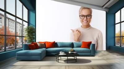 Cose-up shot of upbeat good-looking cheerful young female in glasses with bun hairstyle having awesome mood pointing left and up with thumb as introducing cool promotion over white wall Wall mural