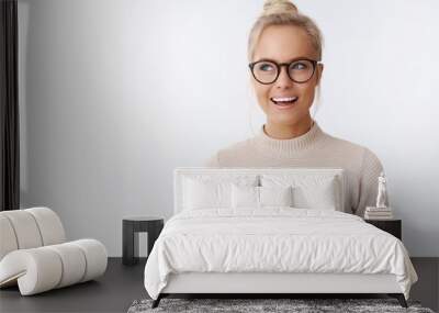 Creative and happy attractive fair-haired blond caucasian female in sweater and glasses having interesting and awesome idea looking at upper left corner daydreaming smiling satisfied over white wall Wall mural