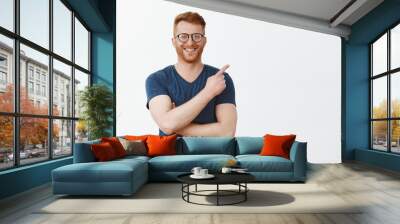 Cute and friendly businessman showing his office and projects to friend. Portrait of handsome redhead male with bristle in glasses and t-shirt, pointing at upper right corner and smiling joyfully Wall mural