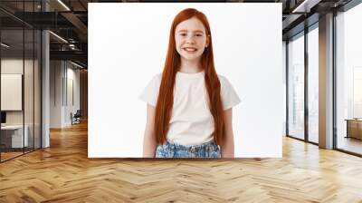 Cute little kid with long red hair smiling and looking happy at camera, standing over white background. Girl child express joy and positive emotions Wall mural