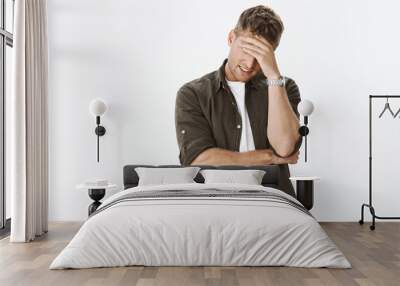 Embarrassed and awkward handsome ex-boyfriend trying hide behind hand as making facepalm gesture so girl would not recognize him, smiling from stress and bother posing against gray background Wall mural