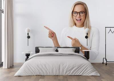 Energized successful good-looking young female entrepreneur in glasses and sweater smiling pointing left amused and entertained feeling excited at awesome course gathering over white wall Wall mural