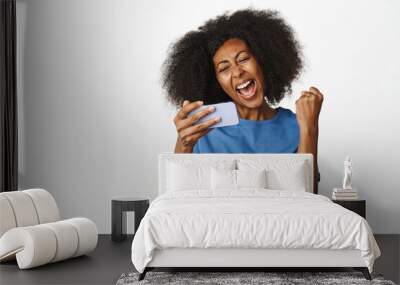 Enthusiastic african woman winning mobile video game. Lady shouts in triumph, celebrating victory on smartphone app, standing in blue t-shirt over white background Wall mural