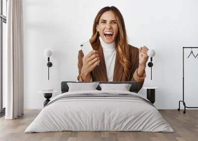 Enthusiastic businesswoman winning on smartphone, achieve goal on mobile phone app and screaming excited, celebrating, standing over white background Wall mural