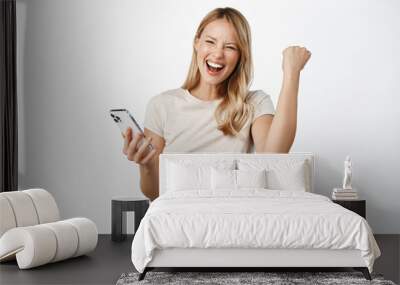 Enthusiastic young woman winning on mobile phone, rejoicing, using smartphone app, celebrating, triumphing on cellphone, white background Wall mural