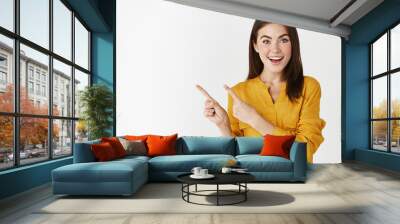 Excited young woman showing banner, pointing fingers left and smiling at camera, standing amazed at white background Wall mural