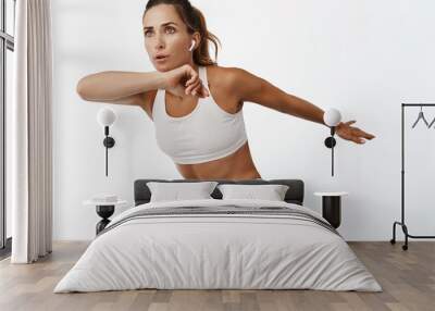 Female athlete doing fitness exercises, control breathing, listening music in wireless headphones, jumping and raising legs, white background Wall mural