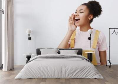 Girl shouting out loud to call friend come downstairs help her. Portrait of charming carefree urban female in yellow trendy overalls, turning left and holding palm near opened mouth, yelling Wall mural