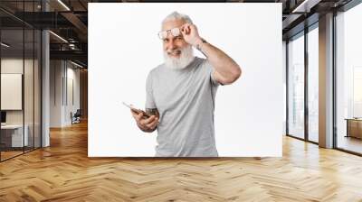 Happy old man with tattoos, take off glasses and using digital tablet, smiling at camera, standing in grey t-shirt against white background Wall mural