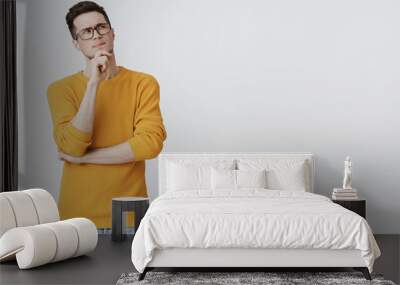 Hmm what if. Smart and thoughtful good-looking guy in geek glasses and cozy yellow sweater holding hand on chin, frowning and looking at upper right corner as thinking, making decision or assumption Wall mural