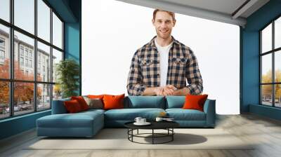 How may I assist you. Smiling pleasant man with red hair, looking content and determined, holding hands together, ready to offer his help, assisting clients, standing over white background Wall mural