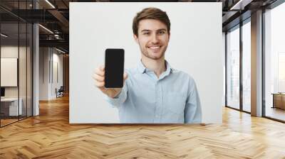 If you seek new phone, buy this model. Portrait of cheerful attractive white male with bristle, pulling hand towards camera and showing smartphone, smiling broadly over gray background Wall mural