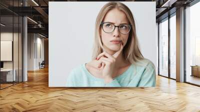 Indoor shot of thoughtful pretty woman has long blonde hair with stylish eyewear, looks aside with pensive expression, plans something on coming weekends, poses against blank wall. Puzzled female Wall mural