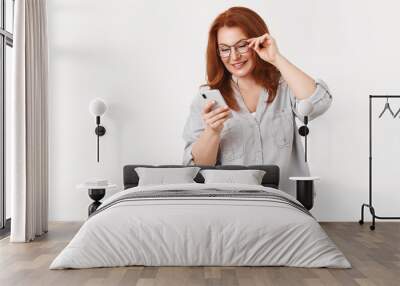 Let look what we got. Gorgeous elegant modern redhead middle-aged mom wear glasses read message check out son send photos social media look smartphone display smiling delighted, white background Wall mural