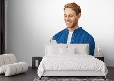 Modern candid guy with phone in hands chatting, message or read screen, smiling at smartphone display while using application, standing over white background Wall mural