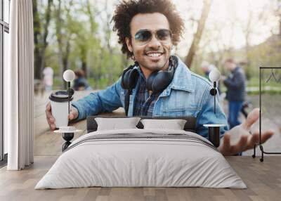 Outdoor shot of fashionable dark-skinned man with afro hairstyle, wearing trendy glasses and headphones over neck, leaning on table in park, drinking coffee and discussing something with gestures. Wall mural