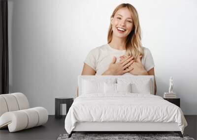 People and faces concept. Young smiling woman holding hands on heart and looking grateful, thank you gesture, feeling tenderness, standing over white background Wall mural