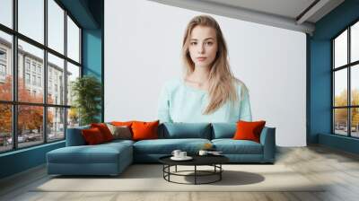 People and lifestyle. Studio shot of attractive young Caucasian dark-eyed female with long dyed blonde hair posing against gray blank wall dressed in casual blue sweater with calm face expression. Wall mural