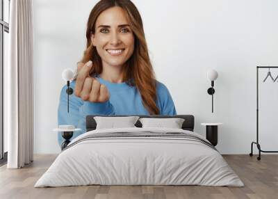 Portrait of beautiful and happy woman smiling, showing finger heart gesture, standing in casual clothes against white background Wall mural