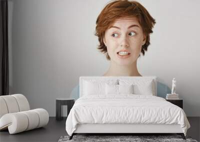 Portrait of confused attractive redhead female feeling awkward, lifting eyebrows and looking aside with weird smile after making mistake and being guilty, saying oops over gray background Wall mural