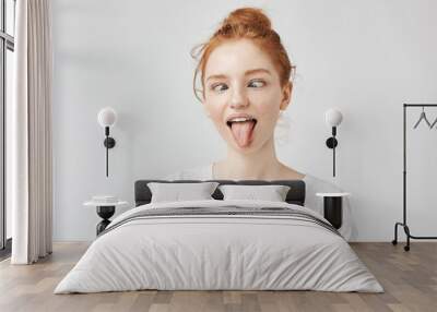 Portrait of crazy and silly yet beautiful freckled girl with eyes crossed, puting out toungue fooling around. Pretending being at audiologist. Easy lifestyle concept on white. Wall mural