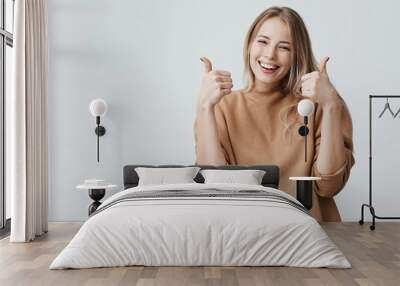 Portrait of fair-haired beautiful female student or customer with broad smile, looking at the camera with happy expression, showing thumbs-up with both hands, achieving study goals. Body language Wall mural